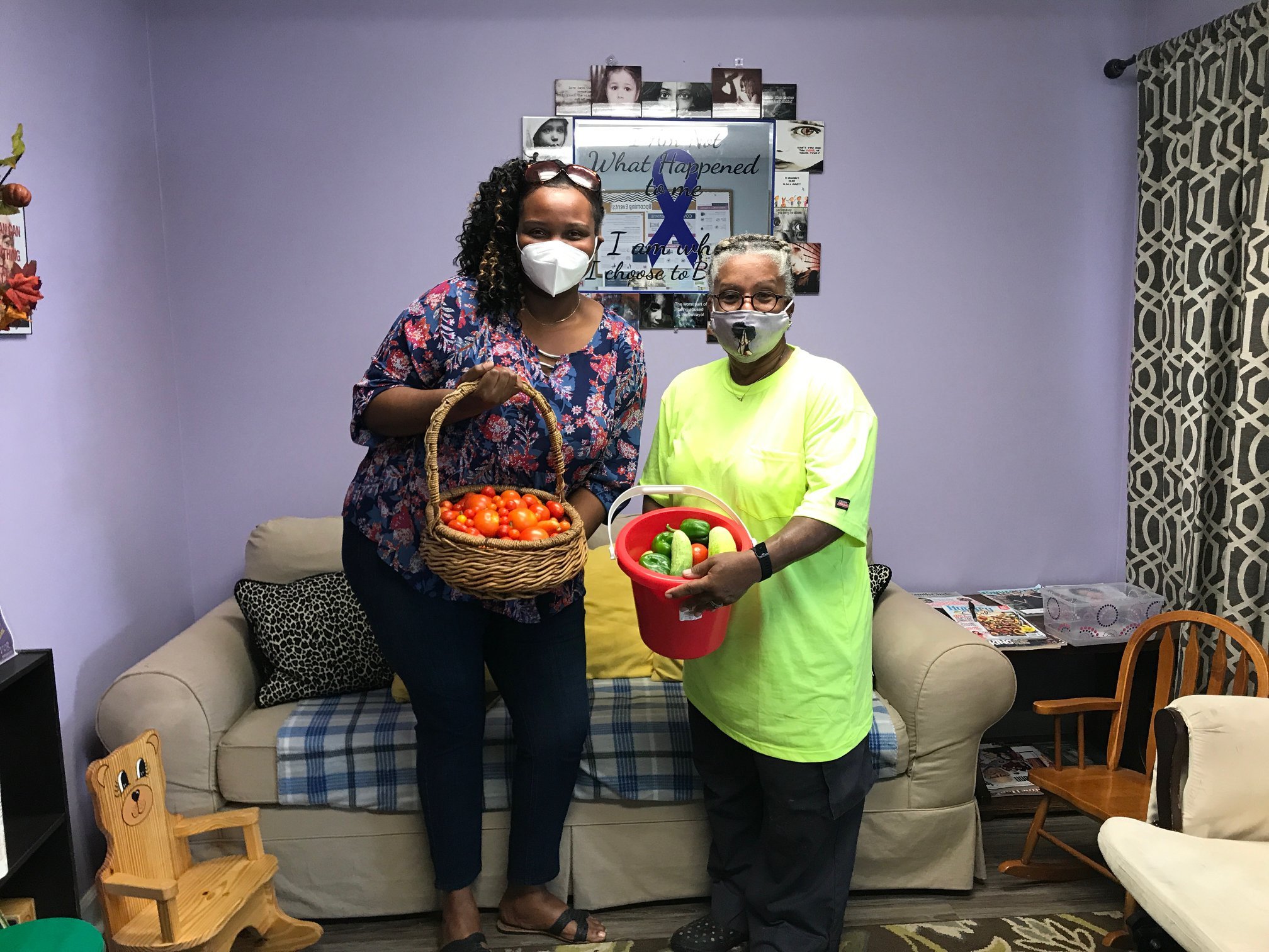Two women wearing masks stand in a cozy living room, one holding a basket of fresh tomatoes and the other a red bucket filled with green peppers and cucumbers. The room features purple walls, a sofa, and decorative items, highlighting a community focus on gardening and healthy eating.