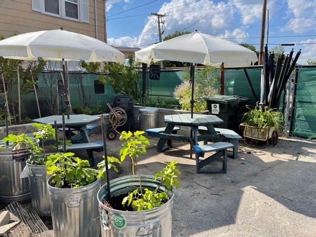 Outdoor seating area featuring picnic tables with umbrellas, surrounded by planters in metal containers, and a green fence in a sunny setting. Ideal for casual gatherings or gardening activities.
