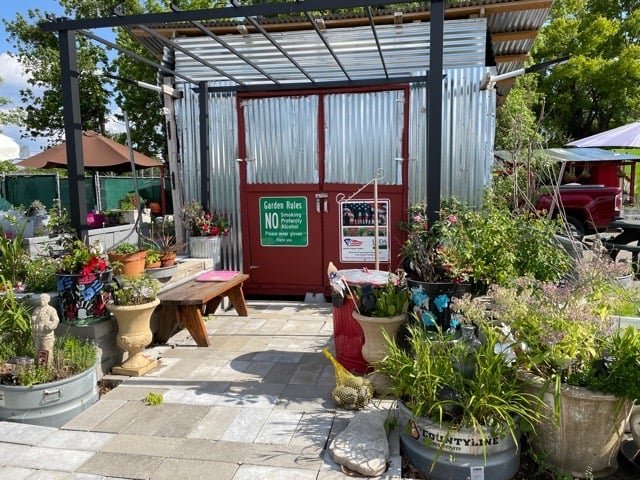 Garden shed entrance surrounded by vibrant flower pots and greenery, featuring a sign with garden rules and a welcoming atmosphere. The area is paved with stone tiles, showcasing a well-maintained community garden space.