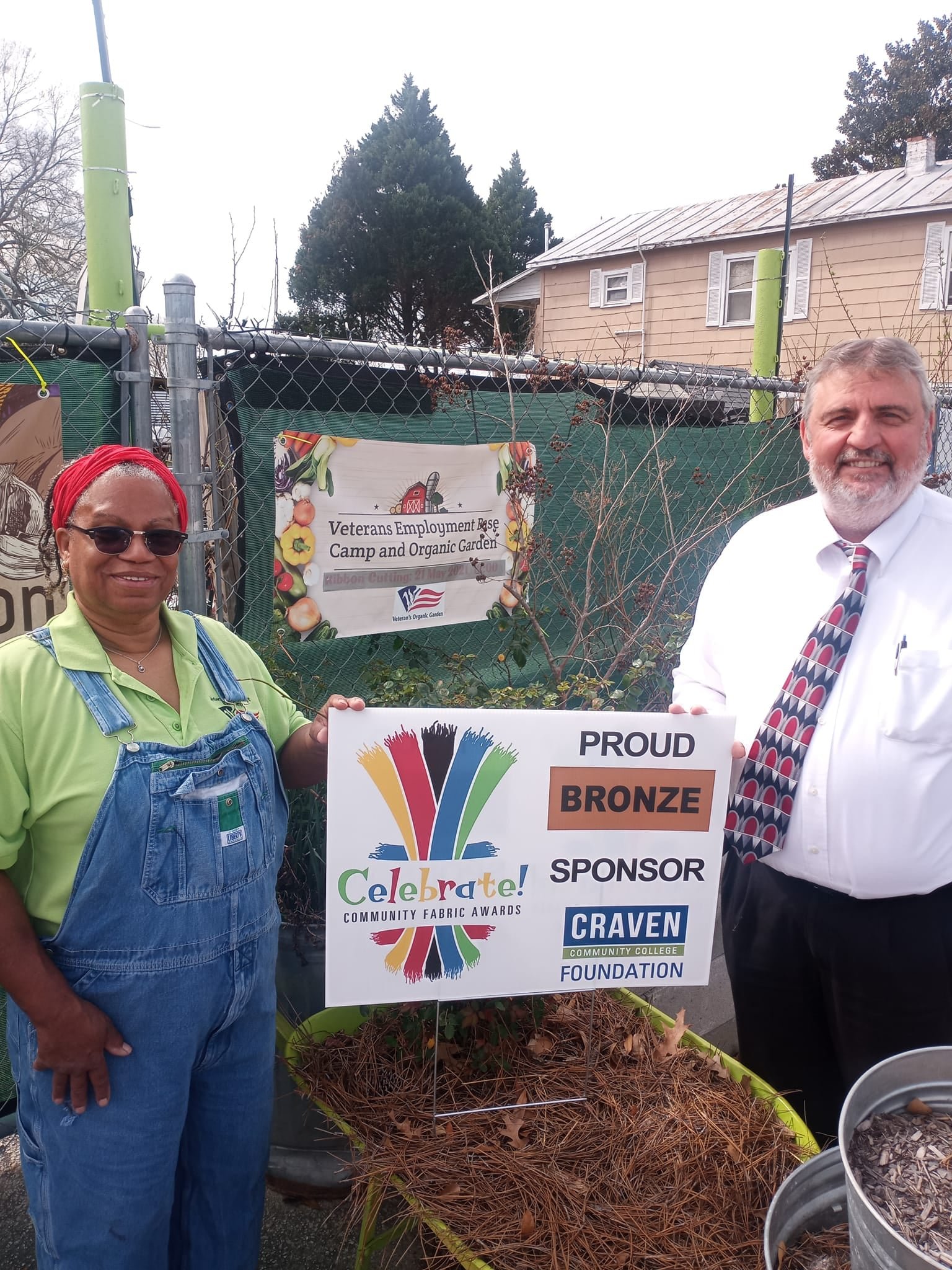 Two individuals stand in front of a community garden, showcasing a sign for the Veterans Employment Age Camp and Organic Garden. One person, wearing overalls and sunglasses, smiles beside a sign indicating the event's sponsorship by the Craven Community College Foundation as a Bronze sponsor for the Celebrate! Community Fabric Awards. The background features a fenced garden area with greenery and a residential building.