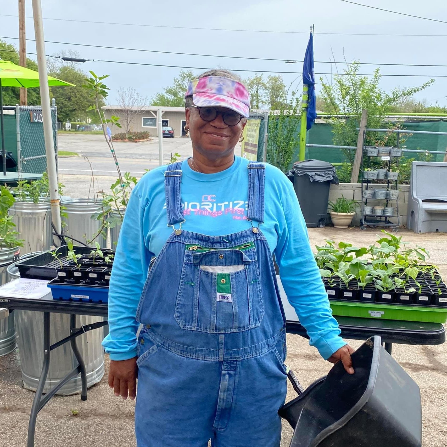 Smiling woman wearing blue overalls and a tie-dye cap stands in a community garden, holding a black container. Green plants are visible on tables in the background, promoting urban gardening and sustainable agriculture.