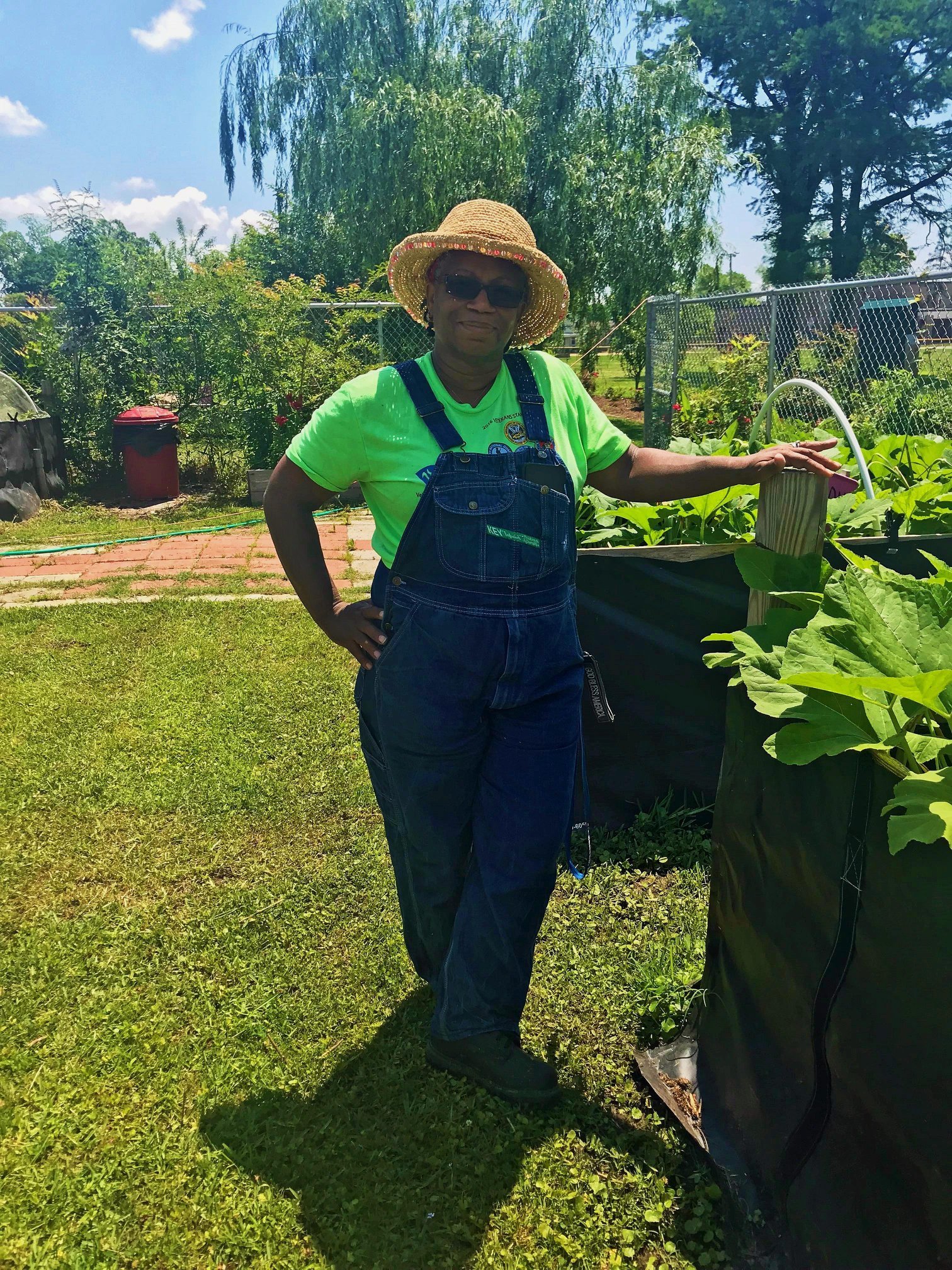 Woman in a straw hat and sunglasses standing in a vegetable garden, wearing a bright green shirt and denim overalls, with lush greenery and raised garden beds in the background under a sunny sky.