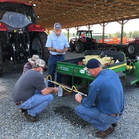 Four farmers inspect and maintain agricultural equipment under a shelter, with tractors parked in the background. They are focused on a green machine, discussing its operation and ensuring proper functionality.
