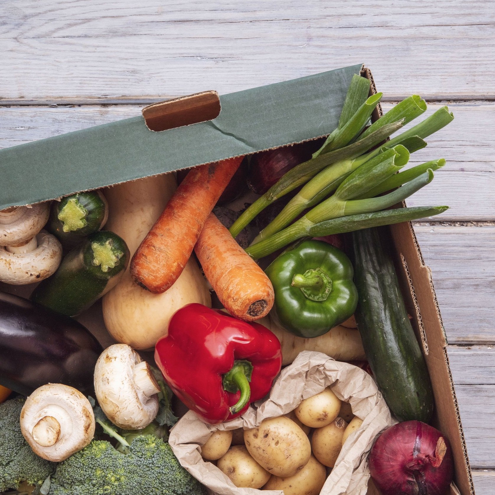 Fresh assorted vegetables in a cardboard box, including carrots, bell peppers, zucchini, mushrooms, potatoes, and green onions, arranged on a wooden surface.