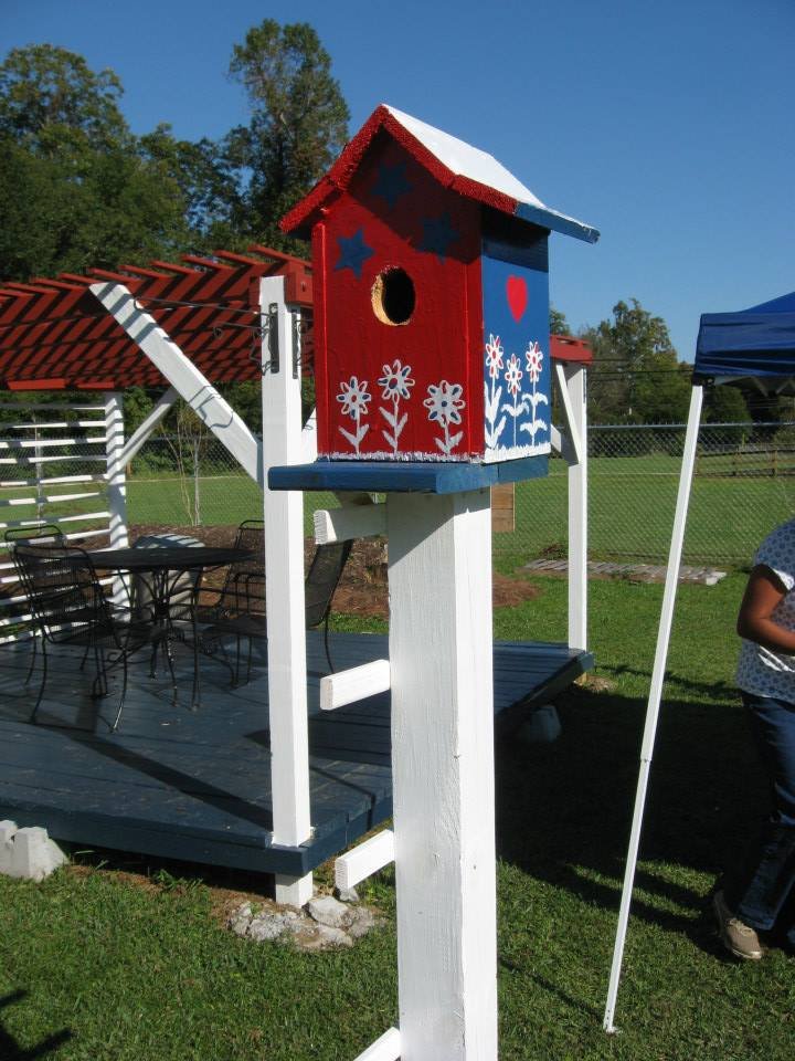 Colorful birdhouse with red and blue painted design, featuring floral patterns and a heart, situated in a sunny outdoor setting with a wooden deck and patio furniture in the background.